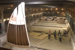 Overhead view of the empty site at Source Park skatepark facility in Hastings, East Sussex. A single modular ramp section is lowered into the subterranean site by crane, whilst four Cambian install contractors in hard hats and highvis jackets walk towards the camera below